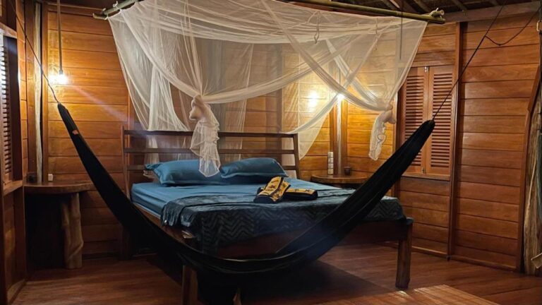Une chambre aux murs en bois éclairée par des lampes, avec un lit sous moustiquaire et un hamac noir