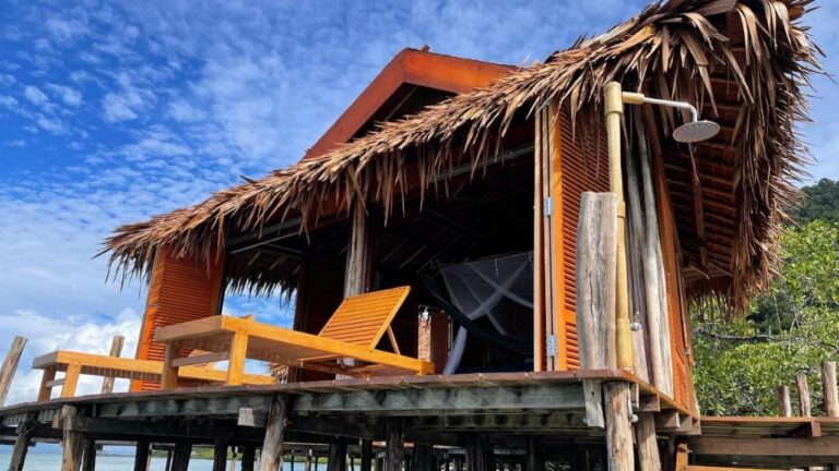 Un bungalow sur pilotis au toit de palmes, avec deux transats en bois et une douche extérieure