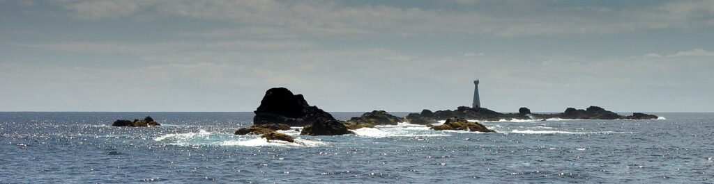 Panorama de la chaîne d'écueils noirs des Formigas à fleur d'eau, ourlés d'écume blanche sur une mer bleue scintillante, avec le phare blanc de Formigão à droite sous un ciel voilé