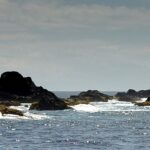 Panorama de la chaîne d'écueils noirs des Formigas à fleur d'eau, ourlés d'écume blanche sur une mer bleue scintillante, avec le phare blanc de Formigão à droite sous un ciel voilé