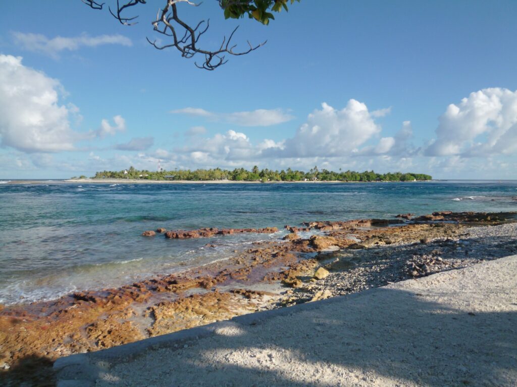 Le chenal de la passe aux eaux agitées, entre un platier de roche rousse au premier plan et un îlot bas couvert de cocotiers sur l'autre rive, sous un ciel de cumulus