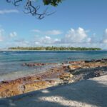 Le chenal de la passe aux eaux agitées, entre un platier de roche rousse au premier plan et un îlot bas couvert de cocotiers sur l'autre rive, sous un ciel de cumulus