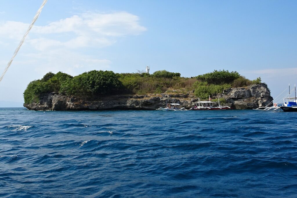 L'île de Pescador vue de la mer, couverte de buissons verts, son phare blanc au sommet et trois bancas de plongée amarrées au pied de la falaise
