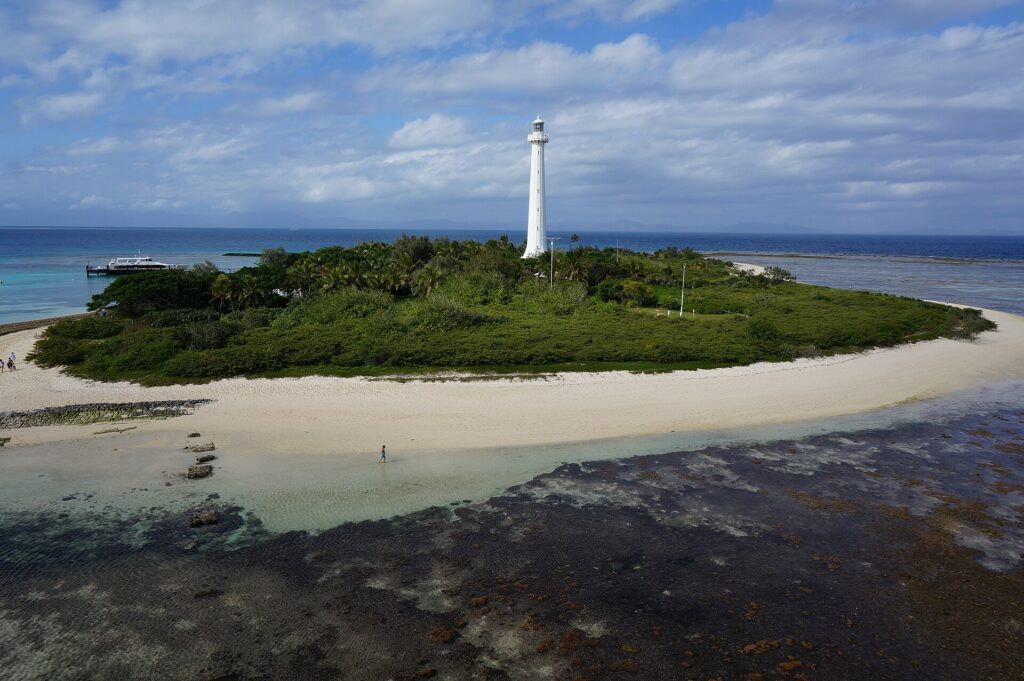 Le grand phare blanc de l'îlot Amédée se dresse au-dessus de la végétation, l'îlot ceinturé de sable blanc, le platier de corail découvert au premier plan et un bateau à quai à gauche