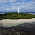 Le grand phare blanc de l'îlot Amédée se dresse au-dessus de la végétation, l'îlot ceinturé de sable blanc, le platier de corail découvert au premier plan et un bateau à quai à gauche
