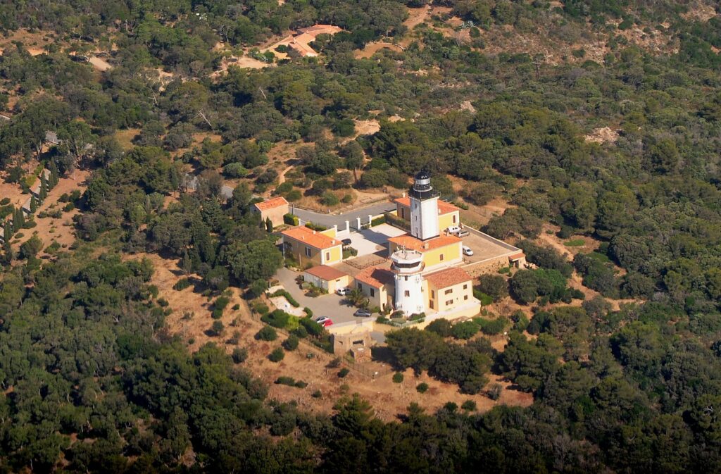 Vue aérienne du phare du cap Camarat, tour blanche à lanterne noire entourée de bâtiments jaunes aux toits de tuiles orange, au milieu d'une forêt méditerranéenne dense.