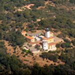 Vue aérienne du phare du cap Camarat, tour blanche à lanterne noire entourée de bâtiments jaunes aux toits de tuiles orange, au milieu d'une forêt méditerranéenne dense.