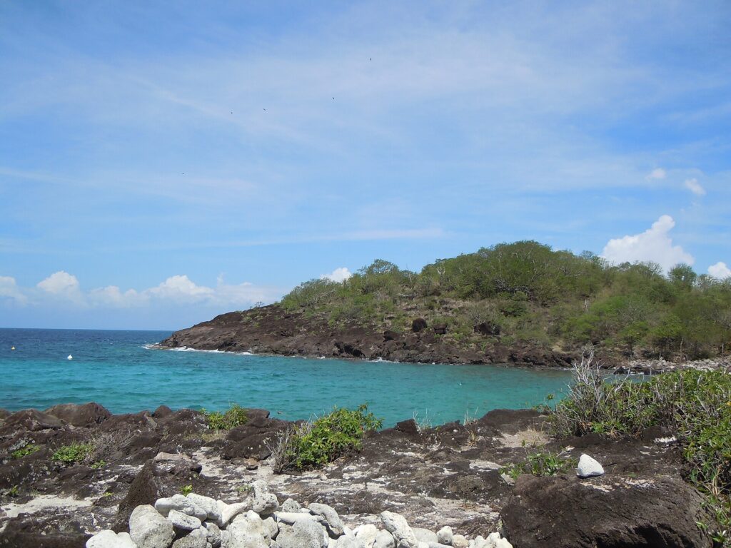 Une anse turquoise abritée par une pointe de roche sombre couverte de broussailles, vue depuis les rochers de l'îlet, sous un ciel bleu