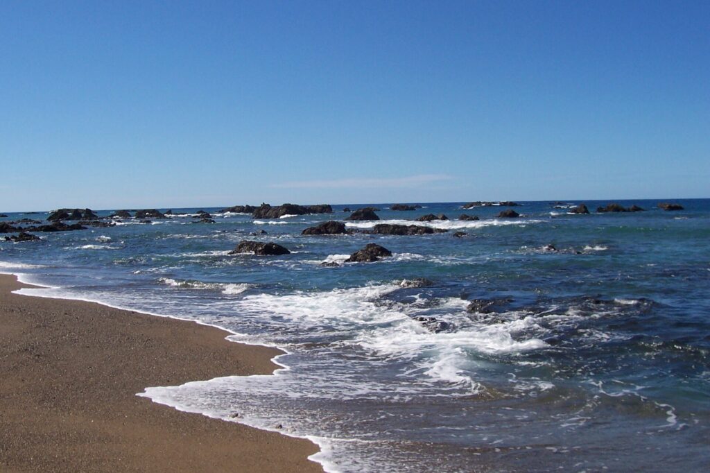 Une plage de sable brun doré où viennent mourir des vagues frangées d'écume, devant une mer bleue parsemée de rochers noirs jusqu'à l'horizon, sous un ciel sans nuage.