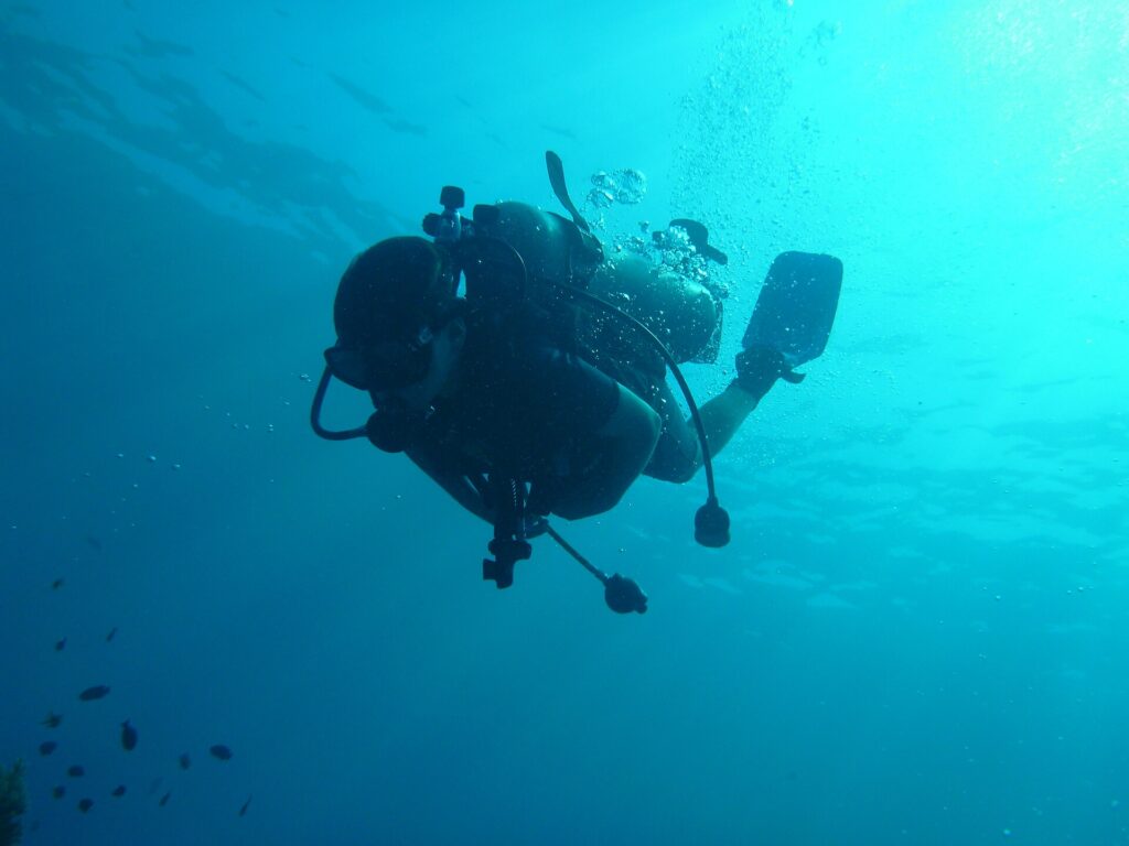 Un plongeur en silhouette vu par en dessous, bouteille, détendeurs et palmes découpés sur l'eau turquoise, ses bulles montant vers la surface éclairée.