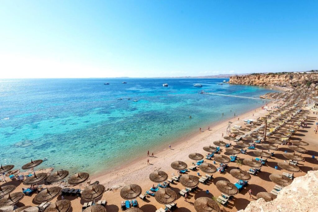 Une longue plage de sable au pied d'une falaise, avec ses parasols de paille et un ponton sur la mer turquoise