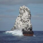 Le pic nord-ouest de Roca Partida, blanc de guano, vu du bateau, une vague qui éclate à sa base et des oiseaux en vol au-dessus