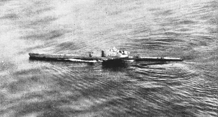 Photo d'archives en noir et blanc, prise d'avion, d'un sous-marin qui file en surface sur une mer ridée, sillage le long de la coque et kiosque au centre.