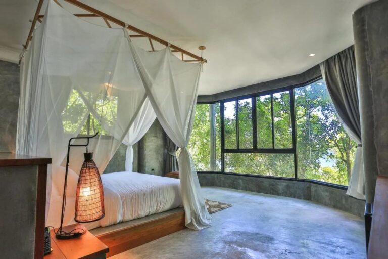 Une chambre aux murs en béton, avec un lit sous moustiquaire et une baie vitrée sur les arbres