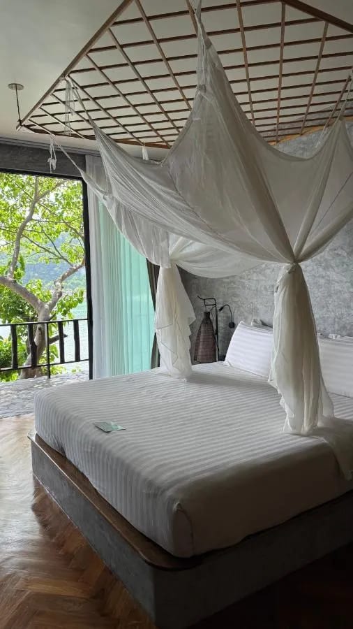 Un lit sous moustiquaire face à une porte vitrée, avec un balcon sur les arbres et la mer