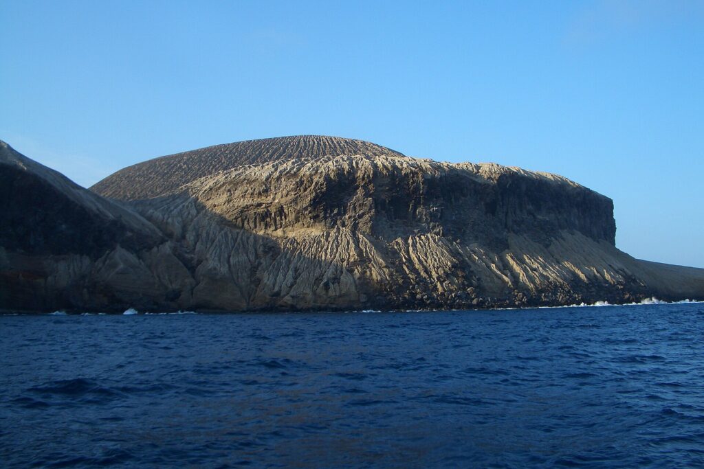 La côte de San Benedicto vue de la mer : le cône de cendres du volcan Bárcena, strié de ravines, au-dessus de falaises sombres et d'une mer bleu foncé