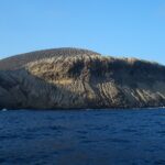 La côte de San Benedicto vue de la mer : le cône de cendres du volcan Bárcena, strié de ravines, au-dessus de falaises sombres et d'une mer bleu foncé