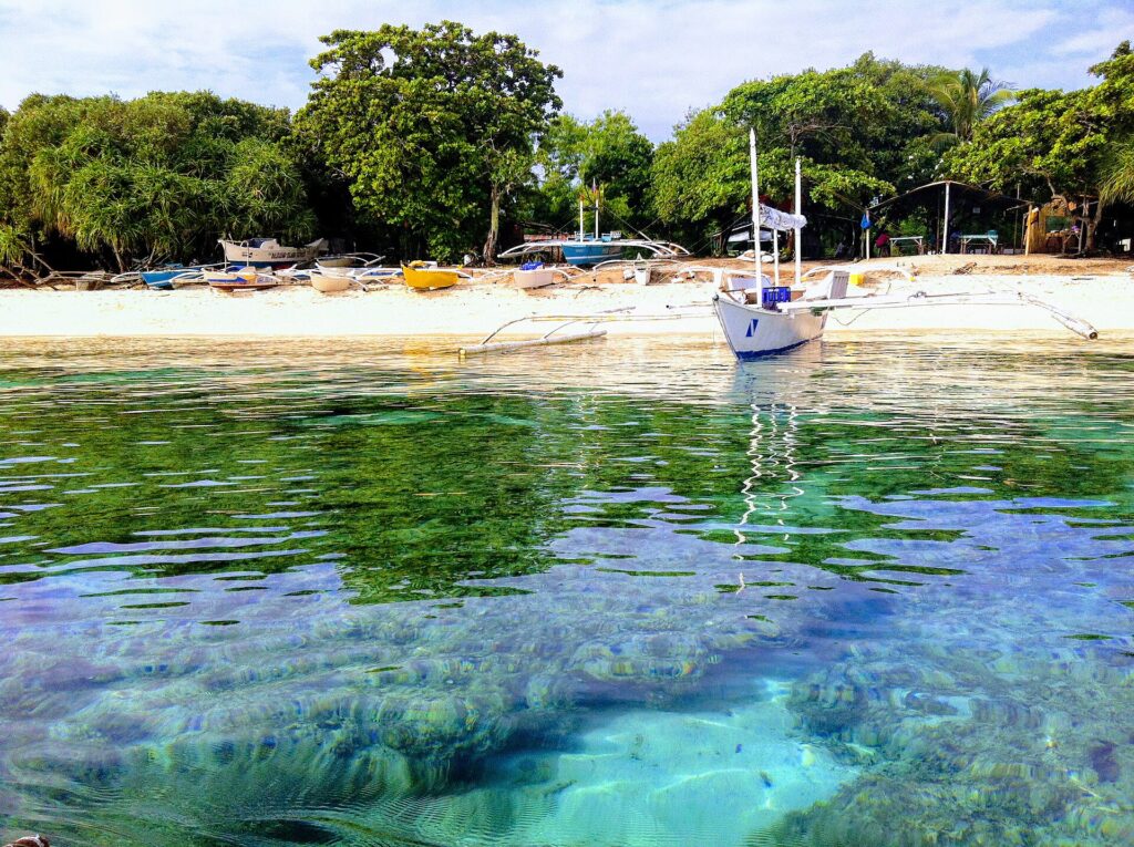 Au ras de l'eau, des patates de corail sous une eau claire et turquoise, devant une plage de sable blanc où mouille une bangka blanche, d'autres barques colorées tirées sous les arbres.
