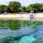 Au ras de l'eau, des patates de corail sous une eau claire et turquoise, devant une plage de sable blanc où mouille une bangka blanche, d'autres barques colorées tirées sous les arbres.