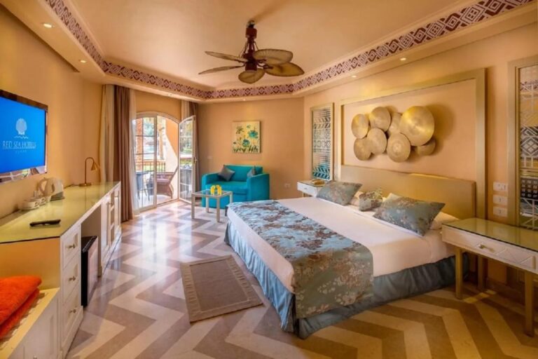 Une chambre au grand lit, avec un ventilateur au plafond, un fauteuil bleu et un sol à chevrons