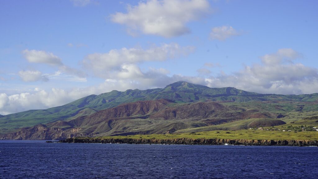 La côte de l'île Socorro vue de la mer : des falaises de lave noire, des collines vertes et rousses et le sommet du volcan dans les nuages