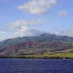 La côte de l'île Socorro vue de la mer : des falaises de lave noire, des collines vertes et rousses et le sommet du volcan dans les nuages