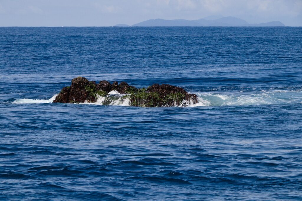 Le sommet de Richelieu Rock émerge à marée basse, rocher sombre couvert d'algues vertes battu par les vagues, les îles Surin à l'horizon