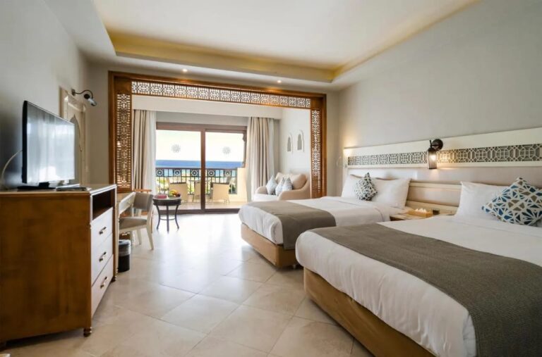Une chambre à deux grands lits, avec un coin salon sous une arche en bois et un balcon face à la mer