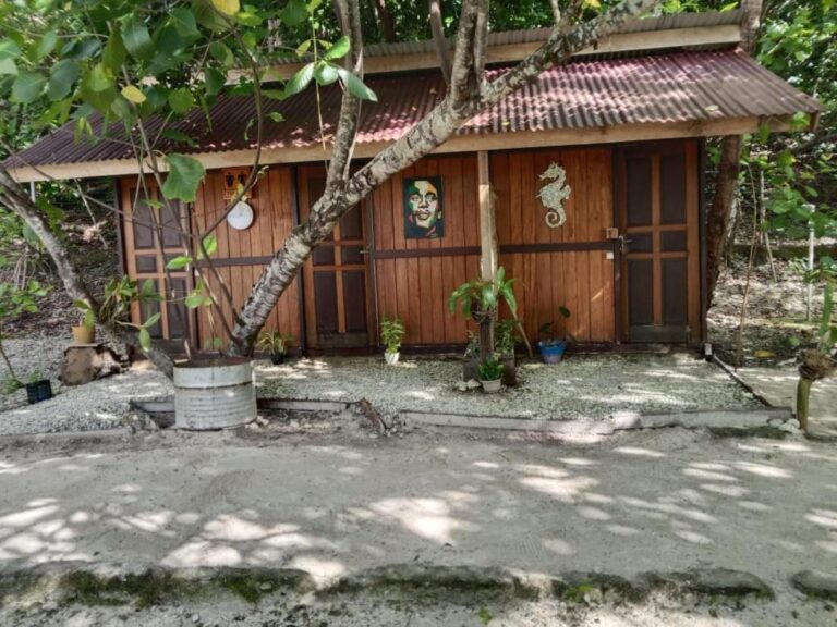 La cabane des toilettes en bois et tôle rouge, au soleil, au bord d'une allée de sable
