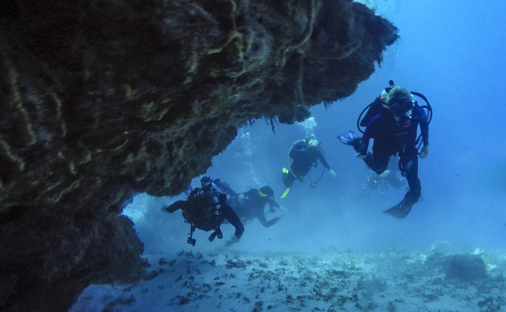 Quatre plongeurs sortent d'un tunnel de corail sur un fond de sable, à Cozumel, le plafond rocheux au-dessus d'eux