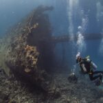 La proue dressée du Giannis D, couverte de concrétions, avec trois plongeurs au pied de l'épave dans une eau bleue chargée de bulles
