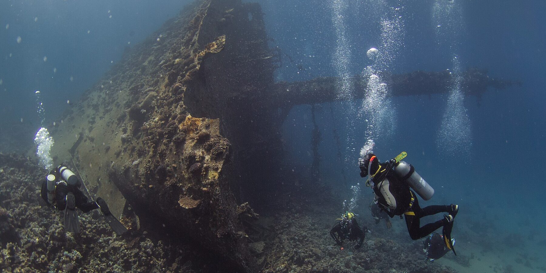 La proue dressée du Giannis D, couverte de concrétions, avec trois plongeurs au pied de l'épave dans une eau bleue chargée de bulles