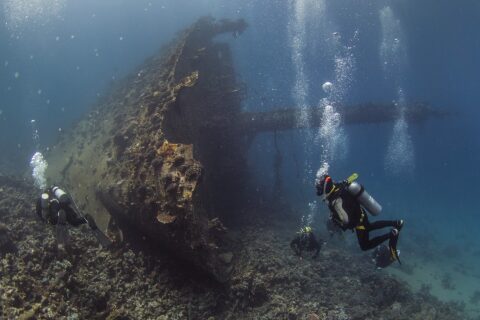La proue dressée du Giannis D, couverte de concrétions, avec trois plongeurs au pied de l'épave dans une eau bleue chargée de bulles