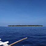 L'île plate et boisée de Balicasag à l'horizon, avec son phare blanc et sa frange de sable clair, vue d'une bangka dont les balanciers blancs occupent le premier plan, sous un ciel bleu sans nuage.