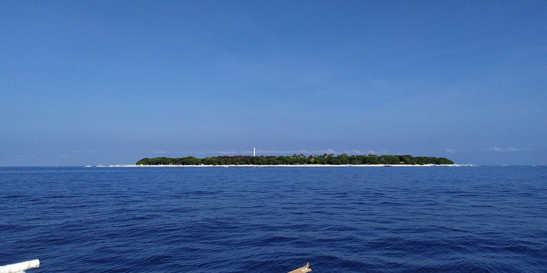L'île plate et boisée de Balicasag à l'horizon, avec son phare blanc et sa frange de sable clair, vue d'une bangka dont les balanciers blancs occupent le premier plan, sous un ciel bleu sans nuage.