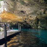 L'œil est du cénote Dos Ojos : une voûte de calcaire hérissée de stalactites au-dessus d'une eau turquoise, un ponton de bois à gauche et des nageurs au fond