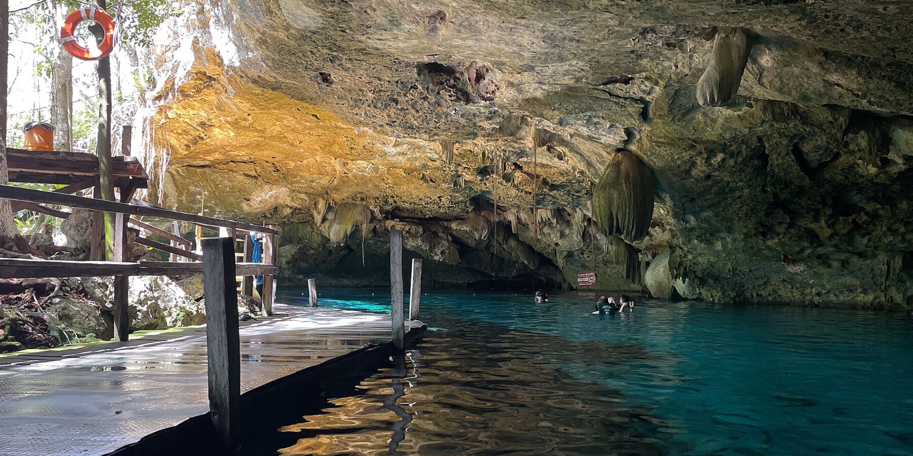 L'œil est du cénote Dos Ojos : une voûte de calcaire hérissée de stalactites au-dessus d'une eau turquoise, un ponton de bois à gauche et des nageurs au fond