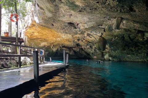 L'œil est du cénote Dos Ojos : une voûte de calcaire hérissée de stalactites au-dessus d'une eau turquoise, un ponton de bois à gauche et des nageurs au fond