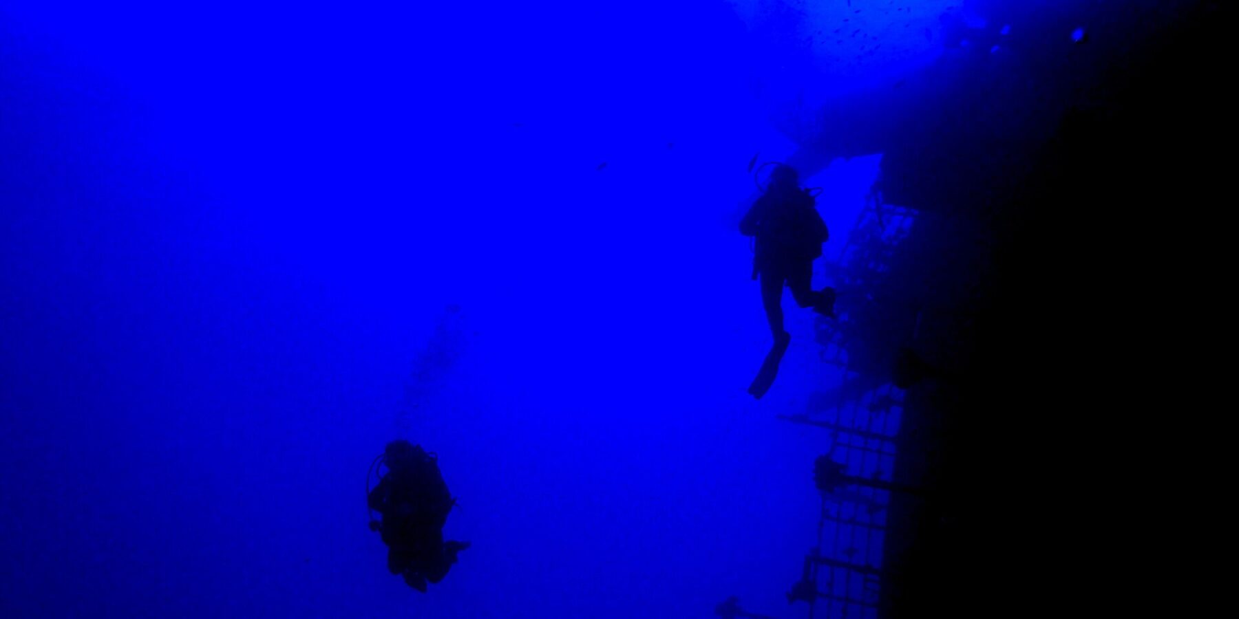 Dans un bleu profond, deux plongeurs en silhouette longent la rambarde de la poupe du Salem Express, le pont incliné à droite