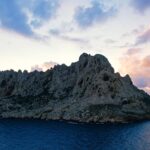 L'île Maïre, masse de calcaire clair aux crêtes déchiquetées, se détache à contre-jour sur un ciel de coucher de soleil aux nuages roses et mauves, au-dessus d'une mer bleu sombre.