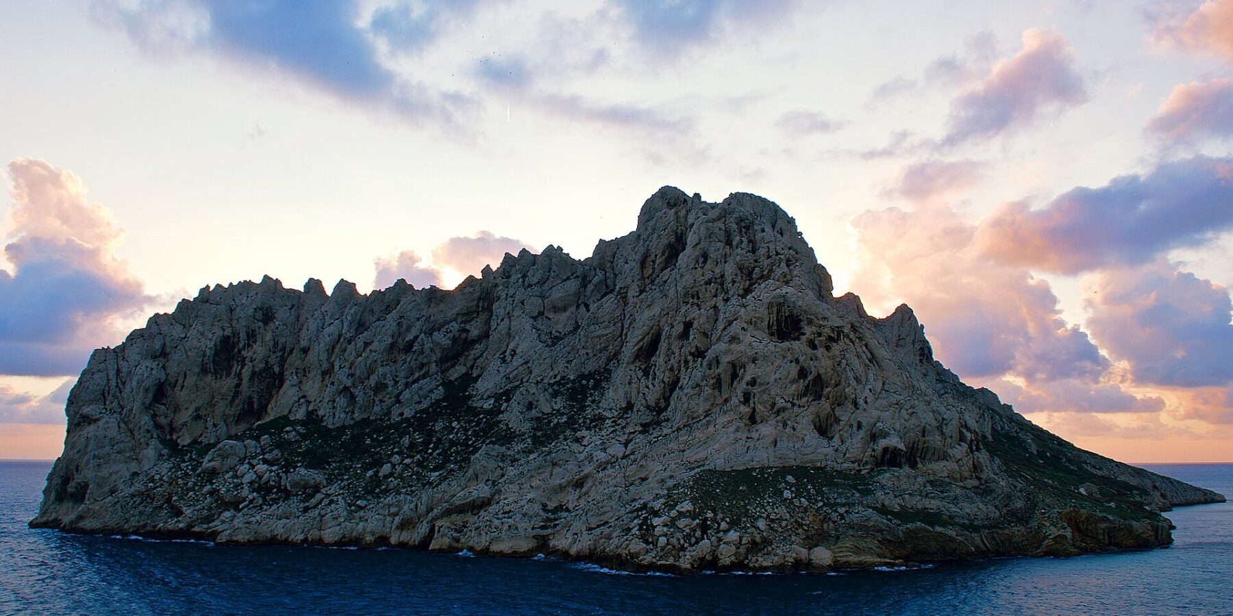 L'île Maïre, masse de calcaire clair aux crêtes déchiquetées, se détache à contre-jour sur un ciel de coucher de soleil aux nuages roses et mauves, au-dessus d'une mer bleu sombre.