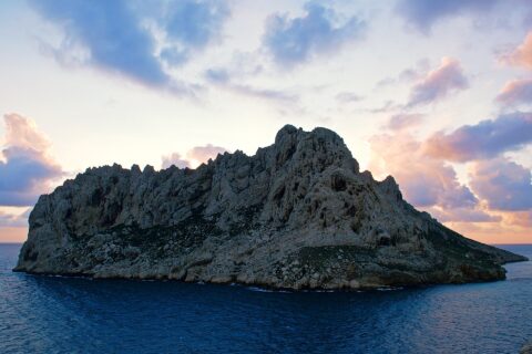 L'île Maïre, masse de calcaire clair aux crêtes déchiquetées, se détache à contre-jour sur un ciel de coucher de soleil aux nuages roses et mauves, au-dessus d'une mer bleu sombre.