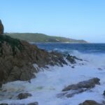Des vagues se brisent en écume blanche sur les rochers de granite de l'Escalet, au premier plan, face à une mer bleu sombre agitée, avec au loin la côte boisée du cap Camarat et son phare blanc.