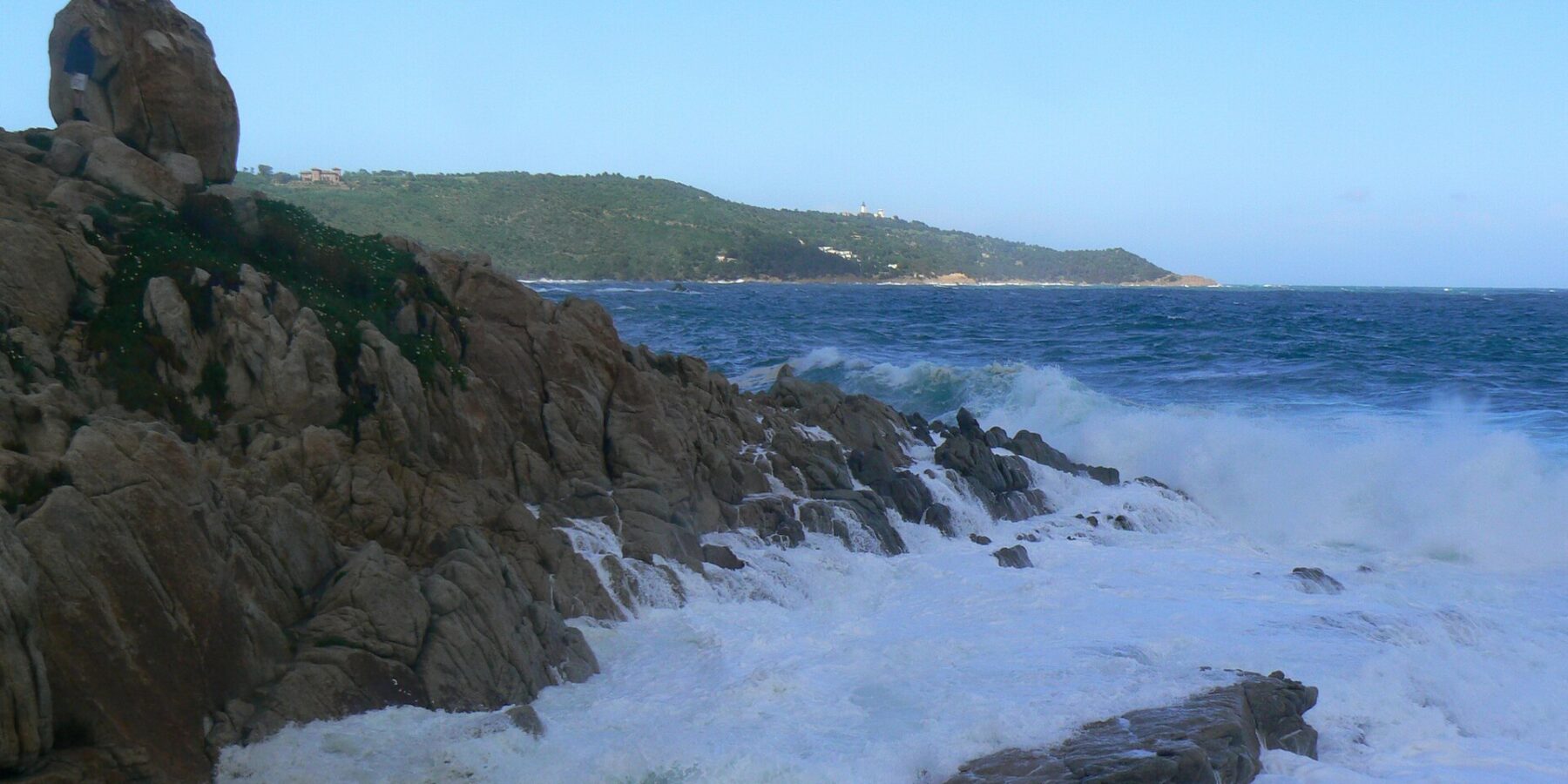 Des vagues se brisent en écume blanche sur les rochers de granite de l'Escalet, au premier plan, face à une mer bleu sombre agitée, avec au loin la côte boisée du cap Camarat et son phare blanc.