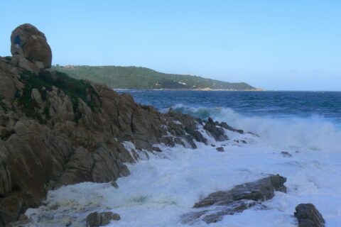 Des vagues se brisent en écume blanche sur les rochers de granite de l'Escalet, au premier plan, face à une mer bleu sombre agitée, avec au loin la côte boisée du cap Camarat et son phare blanc.