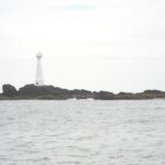 Un phare blanc effilé à porte rouge se dresse sur l'îlot bas et rocheux de Formigão, roches sombres frangées d'algues brunes, au-dessus d'une mer grise et agitée sous un ciel couvert