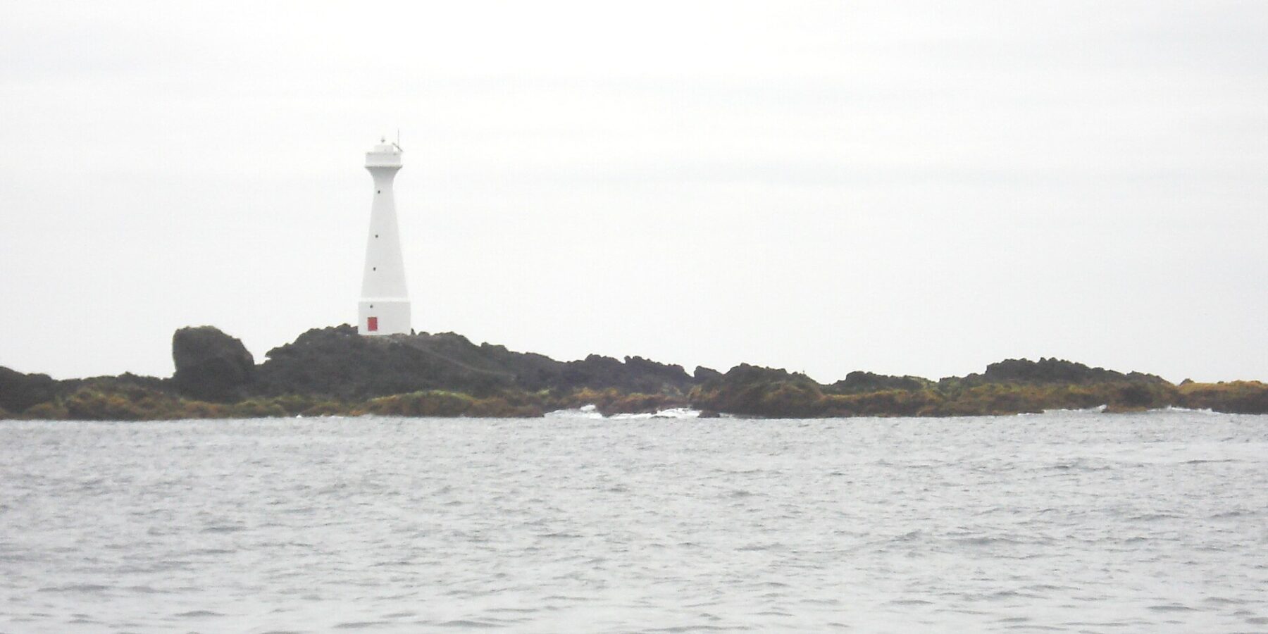 Un phare blanc effilé à porte rouge se dresse sur l'îlot bas et rocheux de Formigão, roches sombres frangées d'algues brunes, au-dessus d'une mer grise et agitée sous un ciel couvert