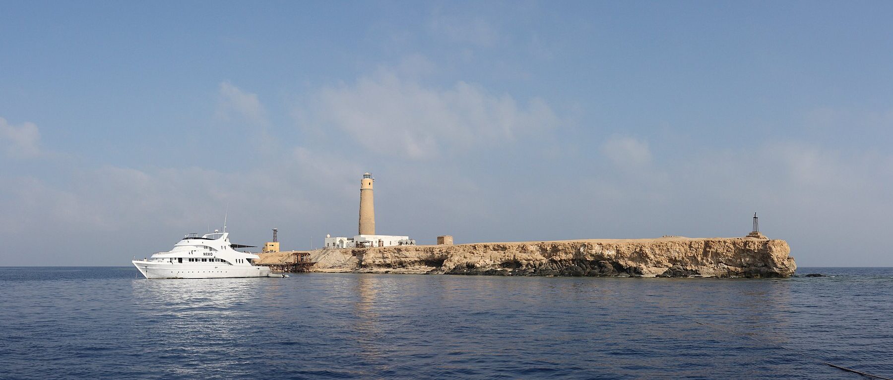 Big Brother vue de la mer : un îlot plat et clair surmonté de son phare, un bateau de croisière blanc amarré devant