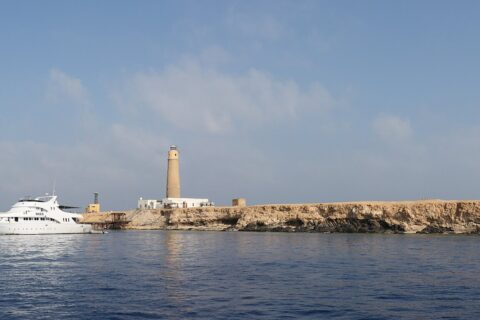 Big Brother vue de la mer : un îlot plat et clair surmonté de son phare, un bateau de croisière blanc amarré devant