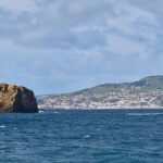 Panorama des deux îlots de tuf ocre de Madalena sur une mer bleu sombre, l'Ilhéu Deitado long et plat à gauche et l'Ilhéu em Pé dressé à droite, avec la ville blanche de Horta et les collines vertes de Faial au fond